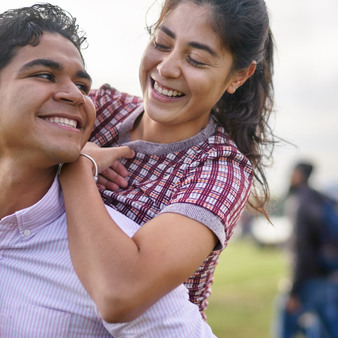 A young man and a young woman who appear to be in their early twenties are smiling at each other as they stand outside.