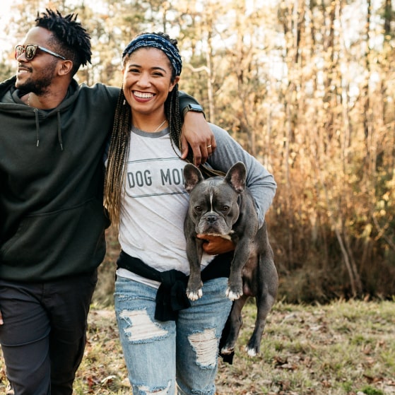 A young woman holding a dog next to a young man are standing in the forest with grass and leaves on the ground.